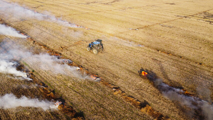 An elevated aerial drone photograph showing a blue agricultural tractor driving across a wide harvested field during controlled burning. © Andrey