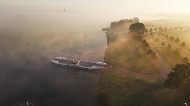Aerial view of a ferry docked on the Meuse river in Kessel, surrounded by morning mist and lush greenery in Kessel, Limburg, Netherlands.