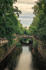 Fototapeta premium Uppsala, Sweden - 20.06.2026: Tranquil urban canal lined with lush green trees and a stone bridge under an overcast sky in Luthagen