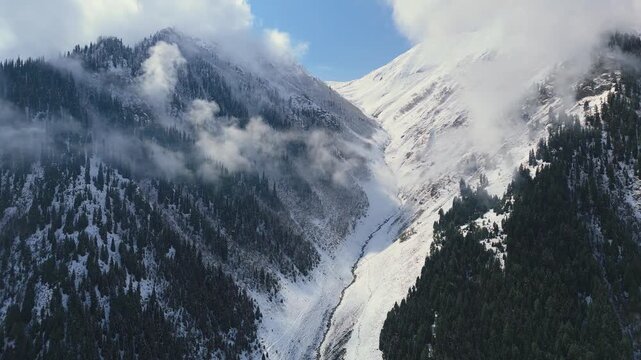 Aerial drone shot of a snow covered mountain chute flanked by pine covered slopes with wispy low hanging clouds