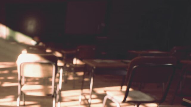 Sunlight streams through windows into a quiet school classroom filled with rows of empty wooden desks and chairs during the late afternoon as dust particles float in the warm glow of the light