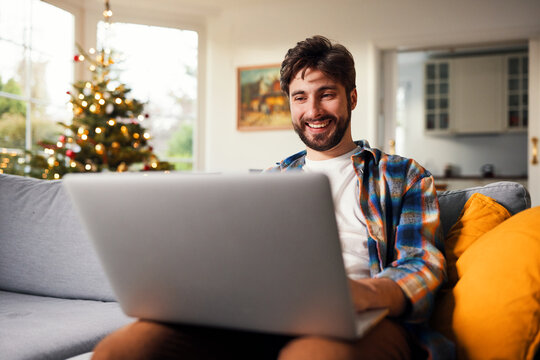 Smiling man using laptop on sofa near christmas tree at home