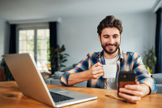 Smiling man with coffee using smartphone at laptop in home office