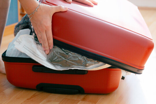 hands pressing on an overpacked red suitcase trying to close it, vacuum bag with clothes sticking out on a wooden floor at home concept of travel, vacation, packing