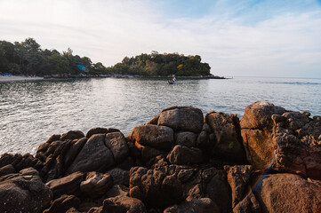 A rocky shoreline frames a calm sea with a tree-covered island and a lone fishing boat near the horizon, warm sunset light and peaceful coastal atmosphere for travel and nature themes. © Loginova