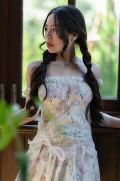 Young Woman with Braided Hair Wearing Floral Dress Near Window in Nature Setting