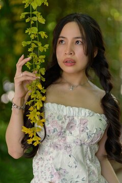 Young Woman with Long Wavy Hair, Wearing Light Dress, Standing Near Green Foliage in Park