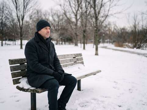 Middle aged man in winter coat and beanie sitting alone on snow covered park bench