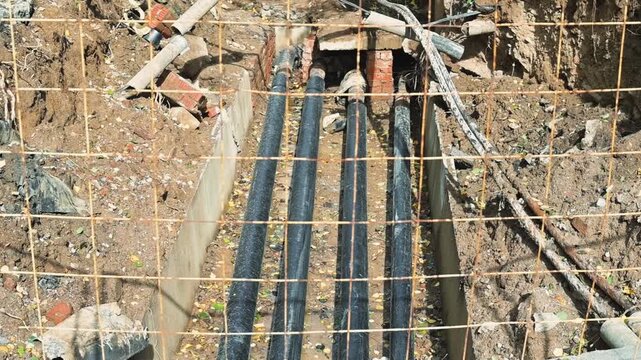 parallel pipes in a trench at an outdoor construction site. The fixed aerial view shows exposed soil, rocks, tools, and equipment around the excavation