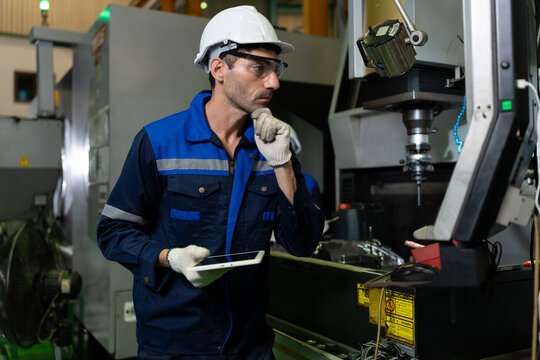 Industrial technician in a blue uniform, hard hat and gloves pauses beside a CNC machine while holding a tablet, suggesting technical assessment, problem solving and precision manufacturing.