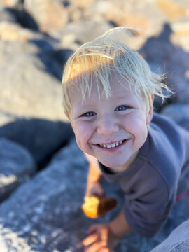 Happy Blond Toddler Boy Eating a Croissant on Rocky Beach