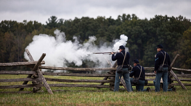Union soldiers in uniform perform a Civil War reenactment, firing muskets behind a split-rail fence in a grassy field with clouds of white gunpowder smoke.