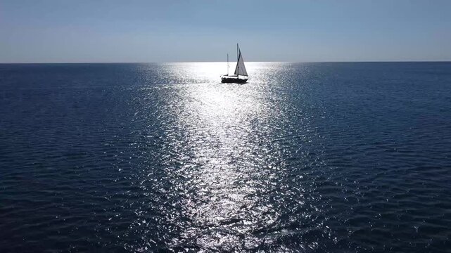 Sailboat cruising on sparkling open water under a bright, sunny sky, aerial drone view. White yacht sailing smoothly across the deep blue sea. Boat navigating calm waters in summer. Sailing vessel.