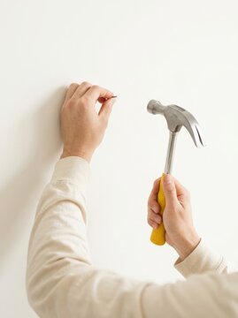 Person hanging small nail on white wall using yellow-handled claw hammer, close-up detail of precise handiwork, indoor DIY repair concept