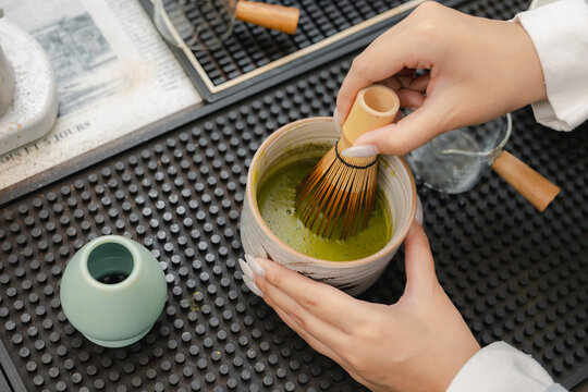 Woman whisking green matcha powder in ceramic cup with calm expression, preparing traditional beverage at cafe counter