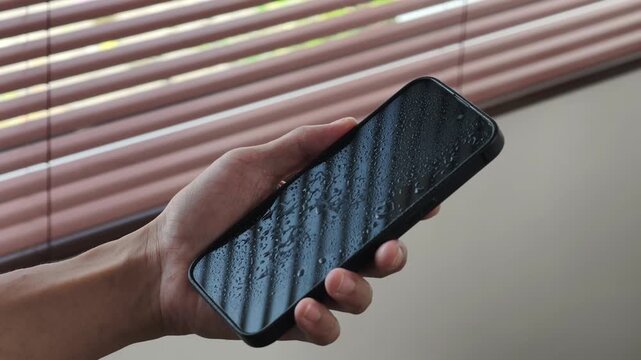 Close-up of hands spraying cleaner onto a black smartphone screen and wiping away moisture with a soft cloth near a window, illustrating electronics care and screen maintenance.