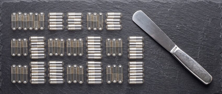 Geometric arrangement of transparent gelatin capsules and metal spatula on dark slate background for pharmaceutical compounding and research
