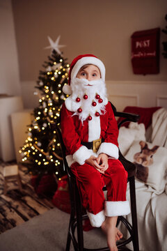 Boy in a Santa Claus costume with a decorated fake beard sitting on a high stool near a Christmas tree