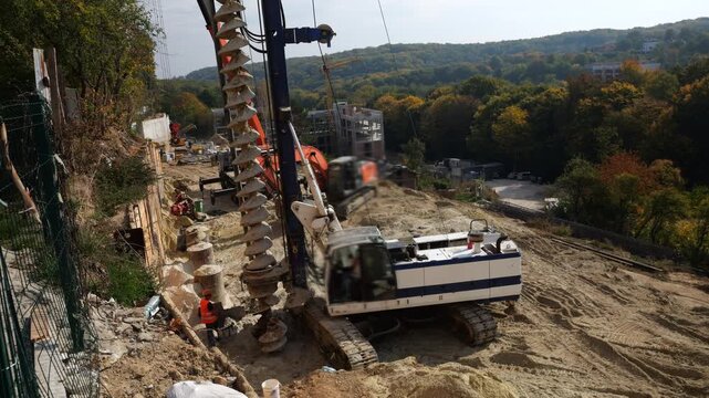 Construction drilling rig with continuous flight auger installs concrete piles for a retaining wall on a steep slope as heavy machinery stabilizes soil at an engineering site.