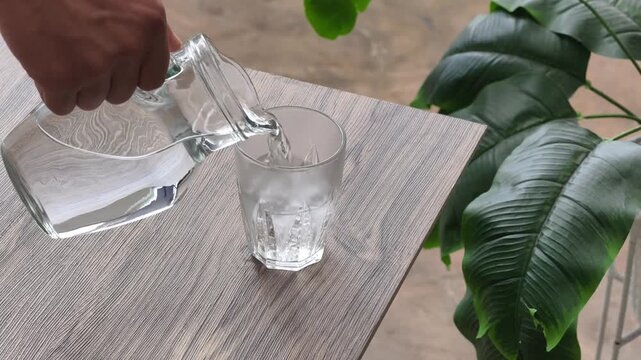 Close-up of a hand pouring clear drinking water from a transparent glass pitcher into a small tumbler on a wooden table, with lush green leaves in the background.