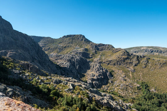 Paisagem montanhosa e rochosa da Serra da Estrela sob um c&eacute;u azul limpo, com encostas de granito, vegeta&ccedil;&atilde;o rasteira e pequenos bosques nos vales em Portugal