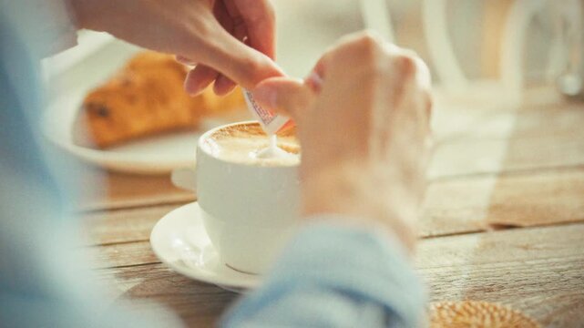 A young woman in light blue adding sugar to her morning cappuccino close-up of hands over a white cup, a fresh croissant, and a wooden table bathed in warm sunlight-a cozy, everyday coffee ritual