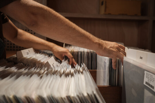 Vinyl records organized by music genre in record store, man browsing LP album sleeves beside Jazz section, searching through analog music collection in wooden display bins.