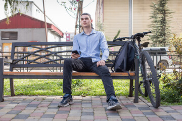 A young man sits on a bench in a park during the day. He looks at his phone while a bicycle rests beside him. © artmim