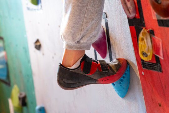 Child's foot grips blue hold. Gray pants, red shoe, white sock. Indoor climbing gym. Focus on grip and footwork