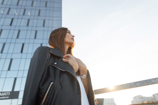 Pensive young woman in a black leather jacket and white shirt, looking up toward a modern glass high-rise. Profile view at sunset with warm sun flares. Low-angle urban portrait.