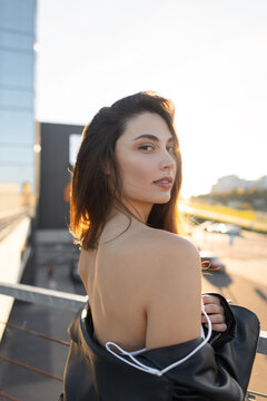 Attractive young woman looking back over her shoulder wearing a white top and black leather jacket on an urban rooftop during golden hour. Backlit by warm sunset light in a modern city