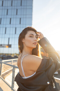 Sensual young woman touching her long hair and looking back on a city rooftop during golden hour, wearing a white top and off-shoulder black leather jacket with a modern building background.