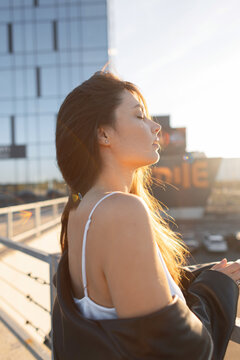 serene young woman with closed eyes on an urban rooftop during a golden hour sunset, backlit by a modern skyscraper. Pretty sexy lady rest at sunset in the city