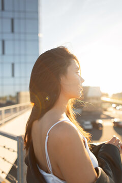 A serene back-lit profile portrait of a young brunette woman in an urban setting at golden hour. She wears a white camisole and holds a leather jacket, gazing at the setting sun over a modern city.