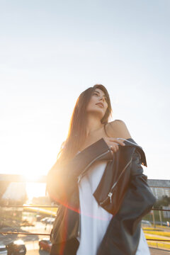 Atmospheric low-angle shot of a young brunette woman on a city bridge at golden hour. Draped in a black leather jacket, looking off-camera. Strong sun flare. Pretty beauty girl at sunset