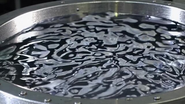 Agitated water swirls across circular stainless steel tank. Reflective liquid develops changing abstract patterns during continuous motion, presenting industrial machinery detail and fluid dynamics.