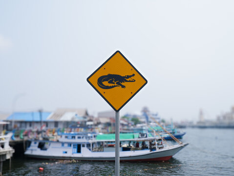 ​Crocodile warning sign near a river harbor with boats in the background, danger caution symbol for wildlife safety
 