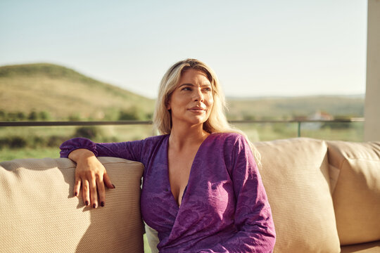 Relaxed Blonde Woman Sitting on Balcony Sofa in Purple Dress During Golden Hour Outdoors