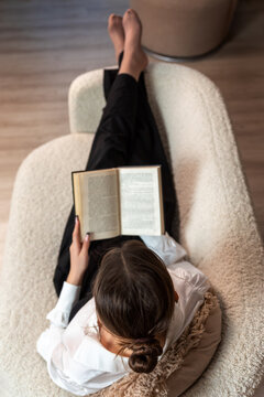 Overhead view of woman in white shirt reading a book on a textured armchair in a relaxed, modern interior.