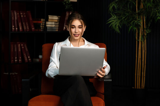 Confident young professional woman working on a laptop in a modern office, surrounded by books and plants. Evening work environment, remote business communication concept.