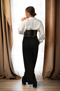 Elegant woman in black trousers and white blouse standing by window, back view. Warm natural light with soft neutral curtains, stylish and contemporary fashion.
