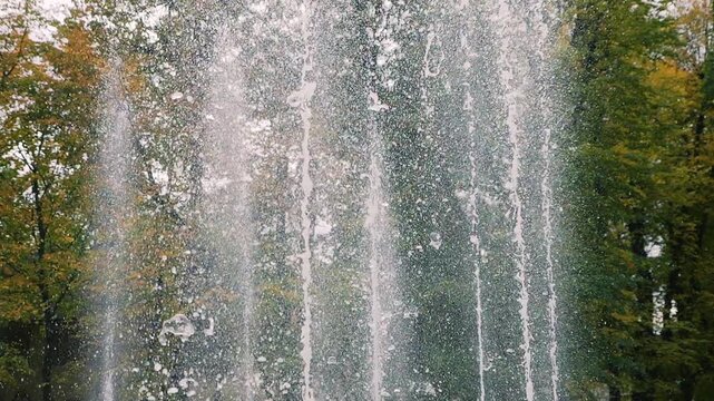 Fountain water jets rise high in slow motion against green trees as crisp splashes create a refreshing liquid wall texture while vertical steady streams spray in the city park.