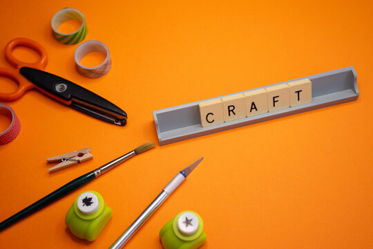Flat lay composition featuring the word -craft- alongside art and stationery supplies, such as scissors, a craft knife, a paintbrush, paper punches, wooden clips, and decorative washi tape on an orang