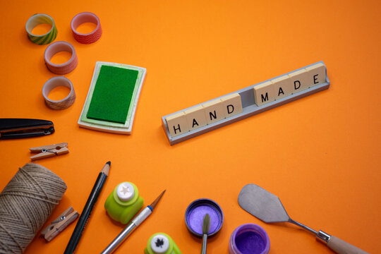 Composition of a flat lay view on an orange background with the phrase 'HAND MADE' formed with tiles made of various materials and crafting tools around it.