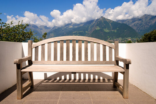 Empty wooden bench on a sunny panoramic terrace overlooking lush Alpine mountains beneath dramatic clouds, evoking tranquility, contemplation, relaxation and scenic escape.