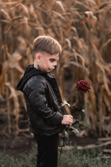 Medium profile portrait of a boy with Halloween skull makeup looking at a dark red rose in a cornfield © Тетяна Хобот