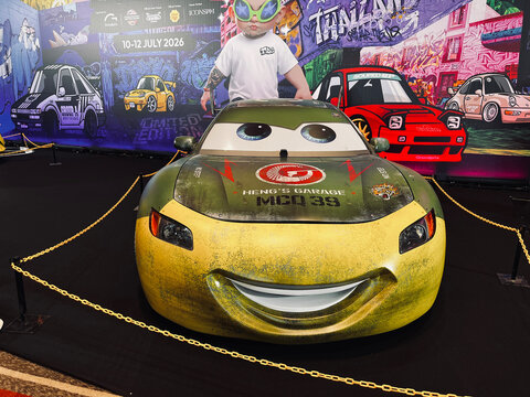 Custom yellow and green Lightning McQueen replica car by Heng's Garage displayed at an automotive and toy event, featuring expressive eyes, racing graphics, and a vibrant motorsport exhibition backdro