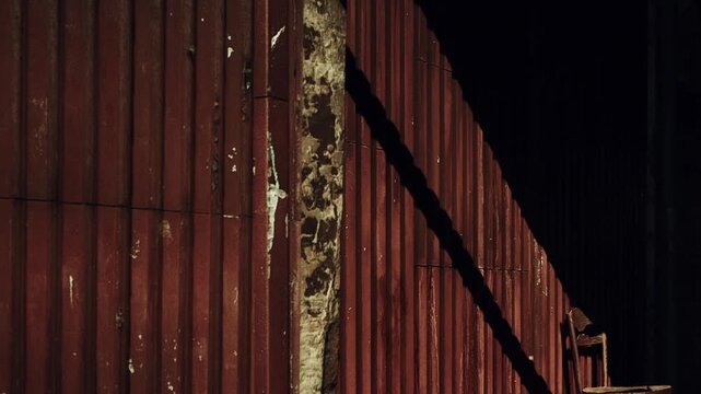 A weathered chair rests against a peeling rust colored wall, casting dramatic shadows in the dim light of a deserted area. The atmosphere feels still and nostalgic, echoing forgotten stories.