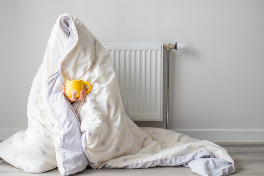 Person wrapped in a blanket sitting beside a radiator with a warm drink. Suitable for heating season and cold weather comfort content