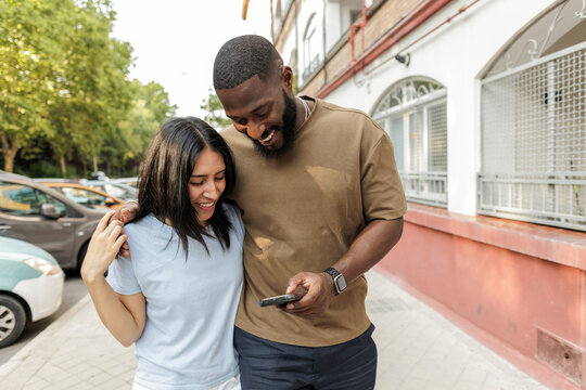 Diverse couple walking street laughing looking at smartphone